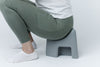 A person wearing olive green leggings and white socks sits on a gray SquatSeat against a clean white studio background.