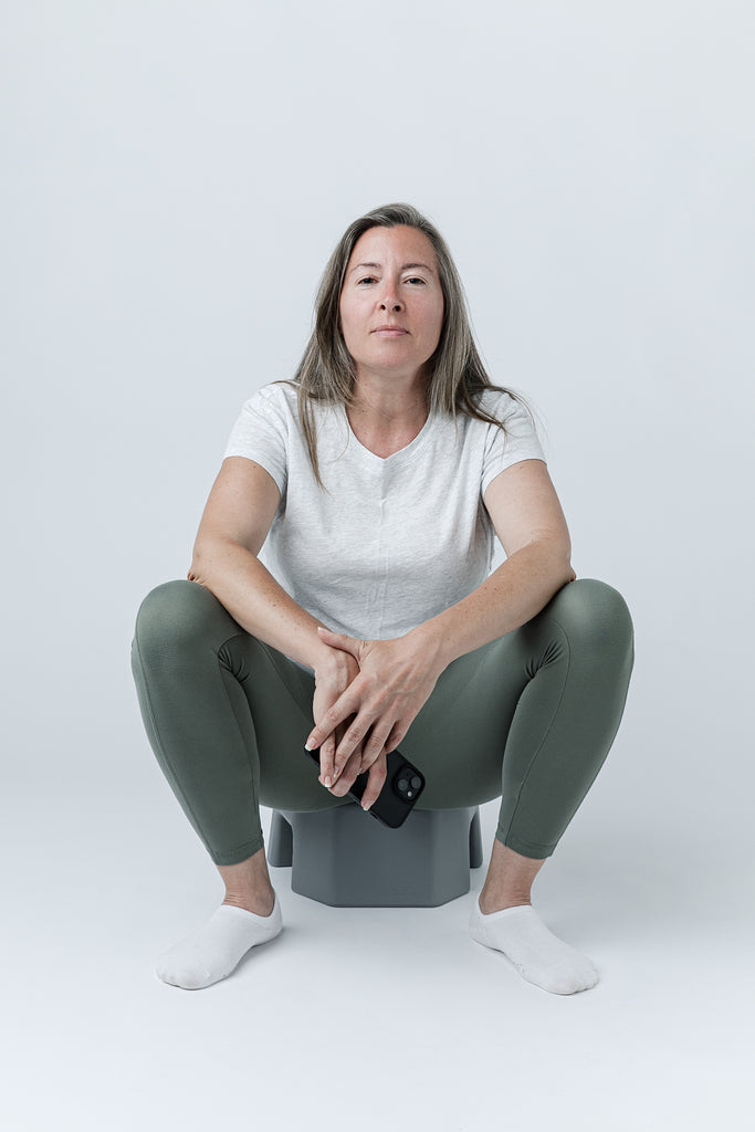 A woman sits in a deep squat on a gray SquatSeat  in a studio setting, wearing a white t-shirt, olive green leggings, and white socks, holding a black smartphone.